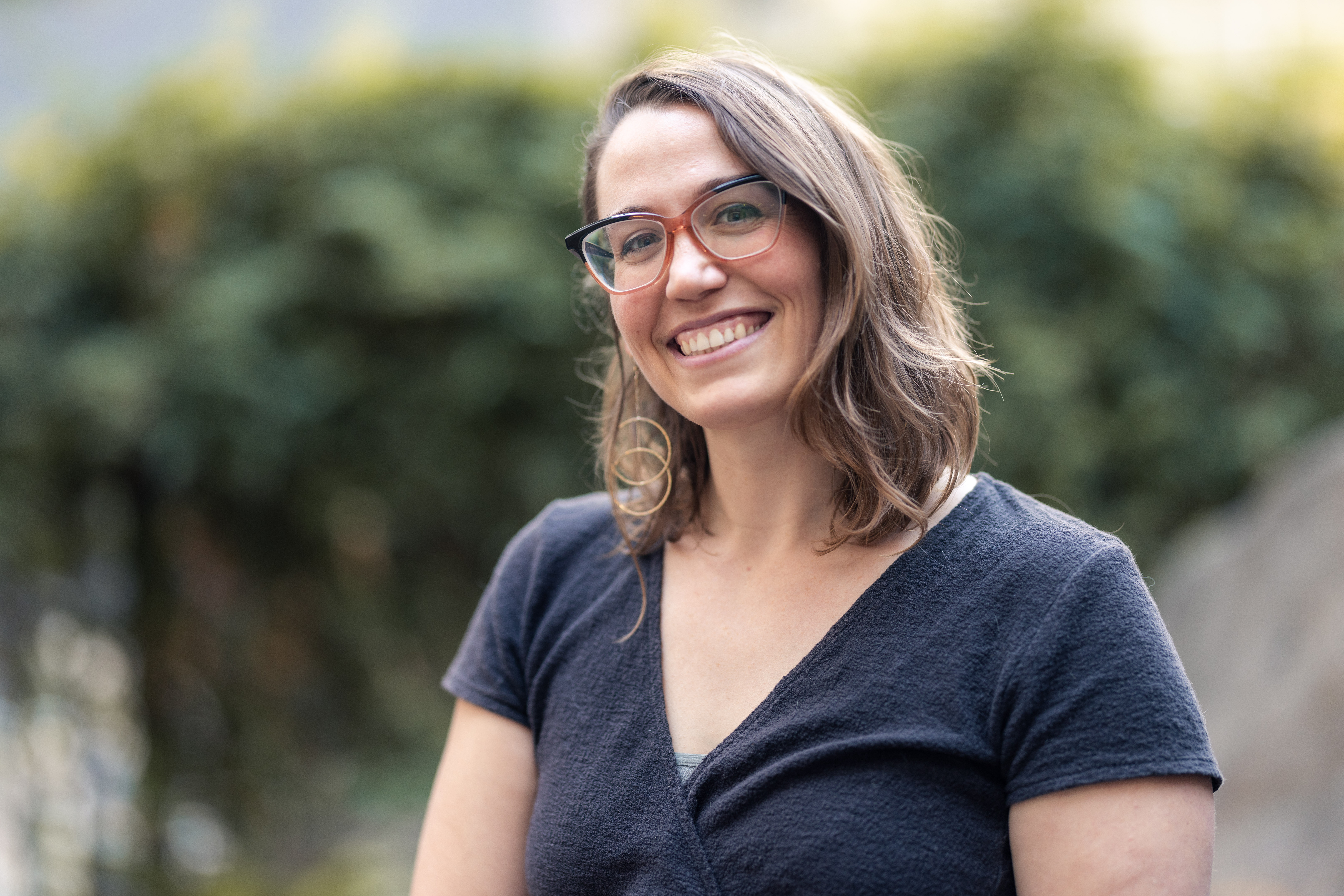 A smiling woman with shoulder-length brown hair wearing glasses and a black top, standing outdoors with greenery in the background.